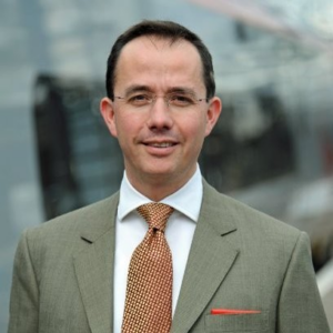 A white man with dark hair, wearing a grey suit and tie, standing outside - the background is blurred