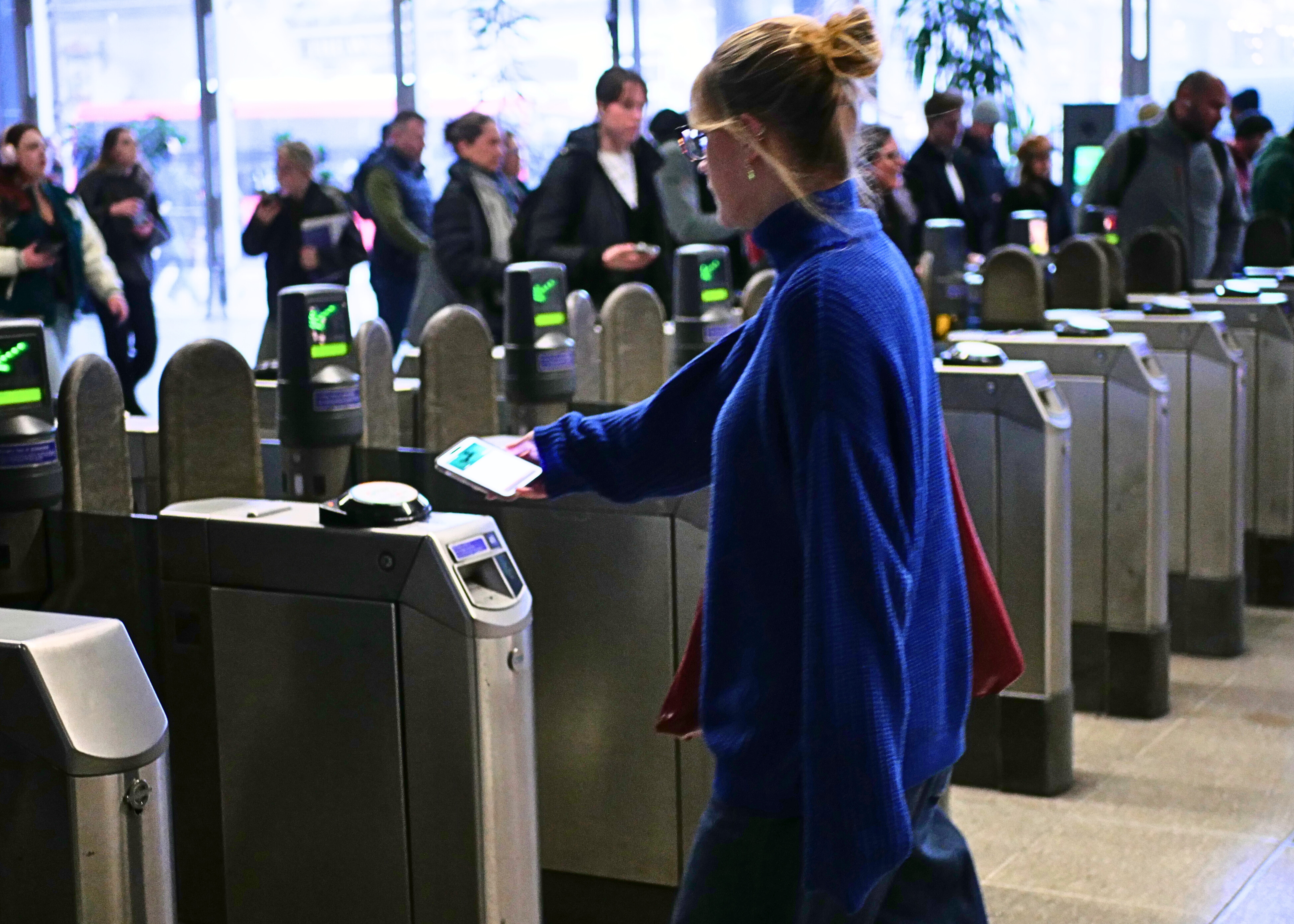 A woman in a blue top using a smartphone to tap through a ticket barrier in a busy station