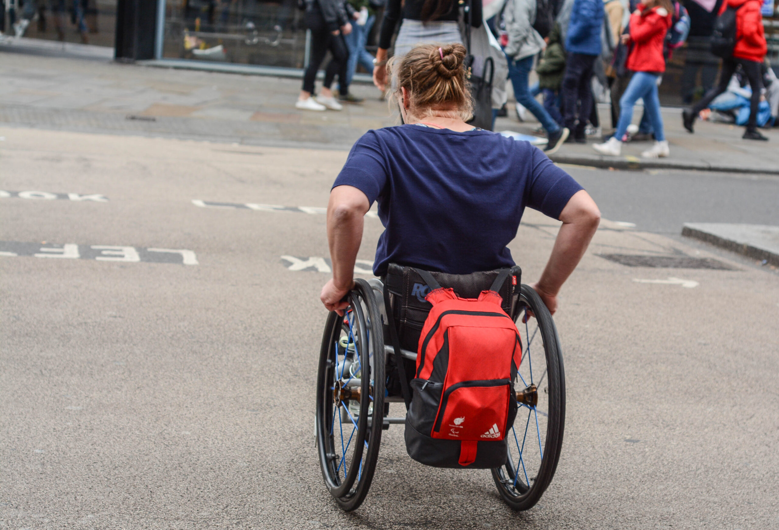 A wheelchair user crossing the road in London. They are wearing a dark blue t-shirt and a red backpack