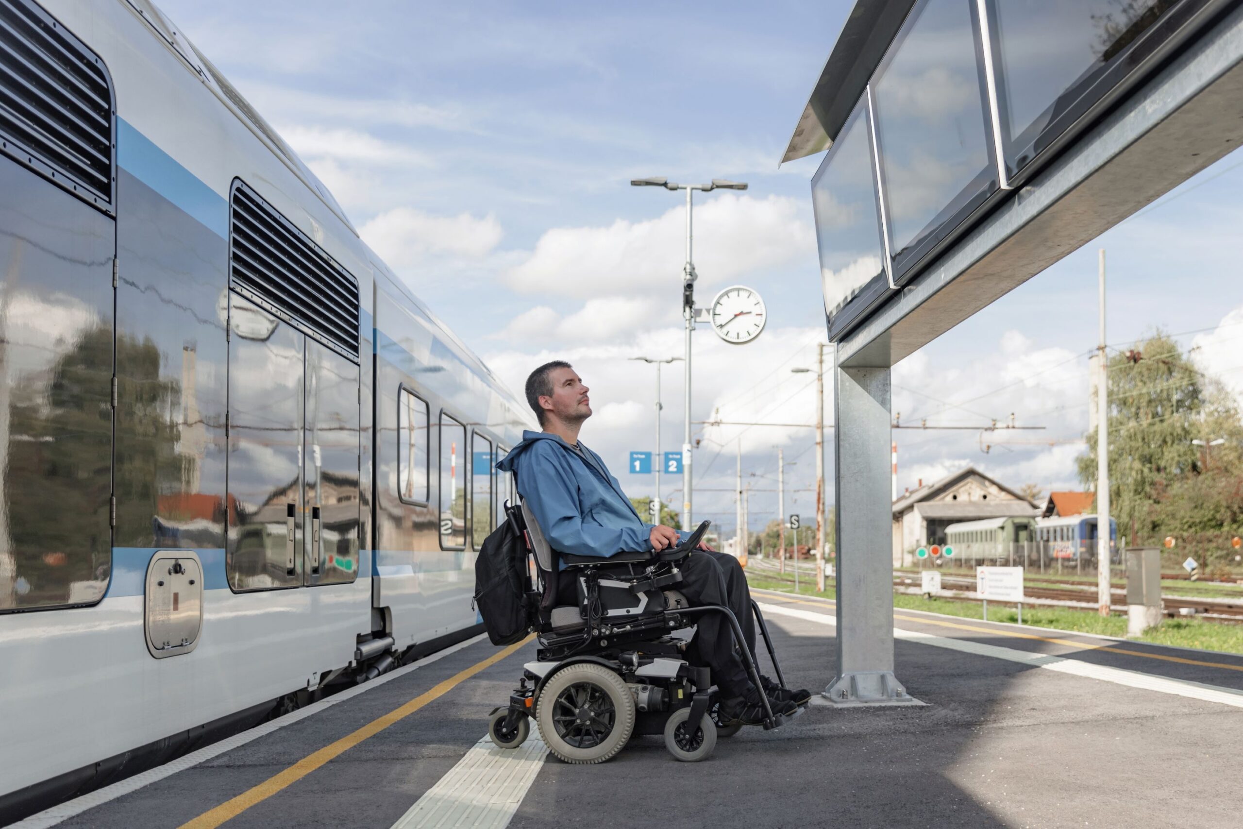 A young white male in a wheelchair at a quiet train platform on a bright day. He is looking at the train departure boards and there is a stationary train behind him