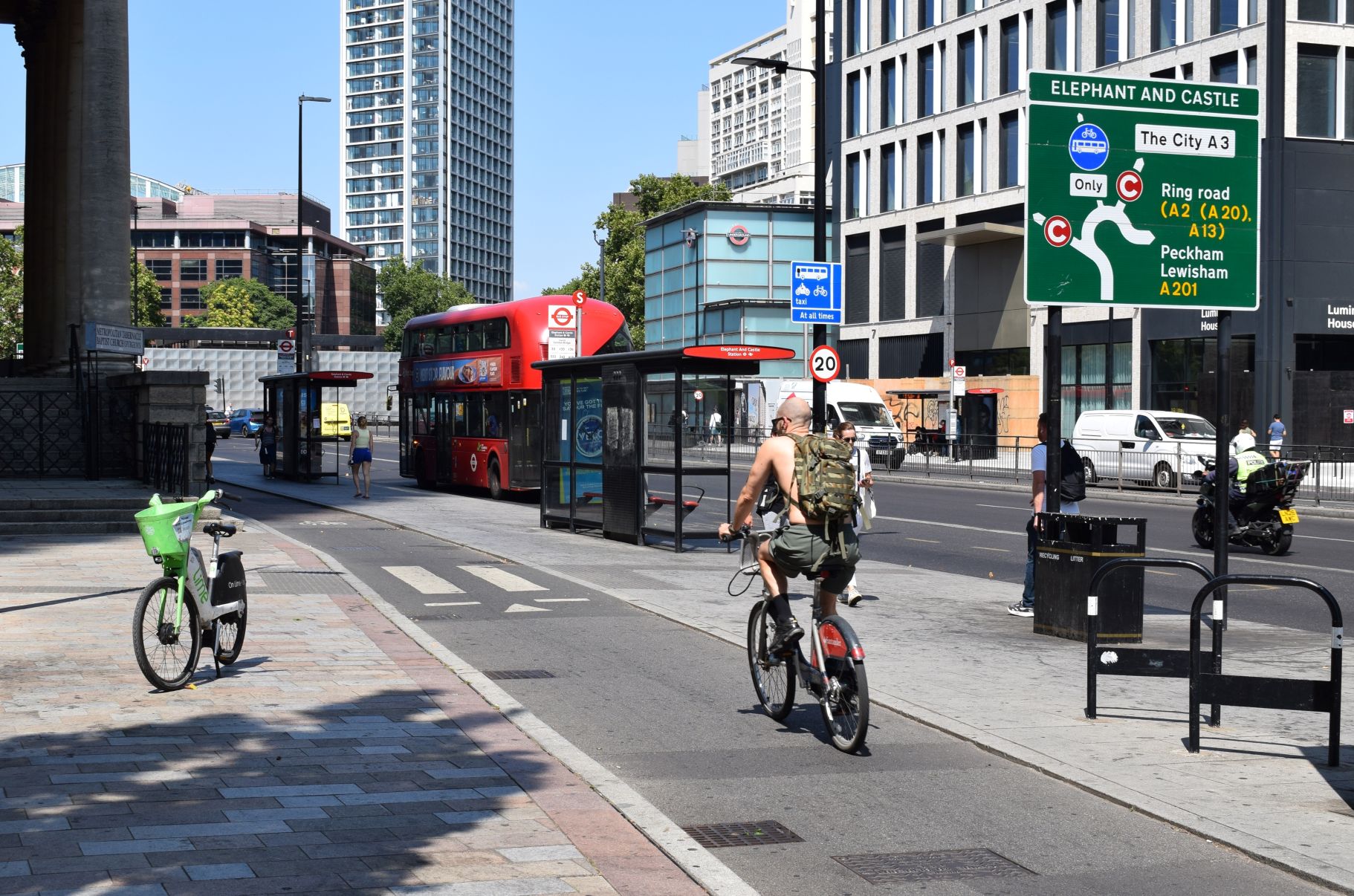 A bus stop bypass on a London street. There is a cycle lane running in-between the pavement and the bus stop. There is a small zebra crossing. A cyclist with their back to us is cycling towards the crossing.